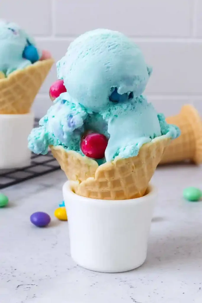 Blue bubble gum ice cream with gumballs in a waffle cone served in a white cup on marble countertop