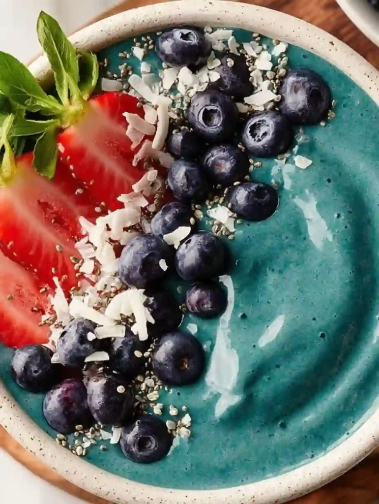 Teal blue spirulina smoothie bowl in stoneware topped with blueberries strawberries coconut flakes and chia seeds
