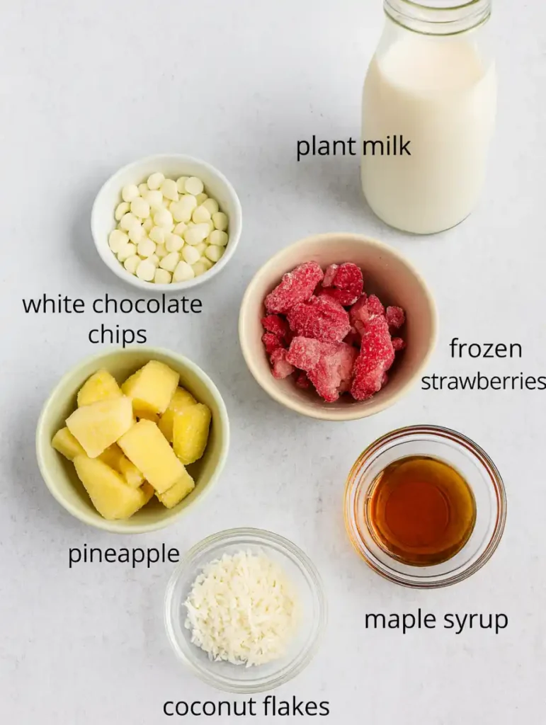 Flat lay of smoothie ingredients including strawberries, pineapple, coconut flakes, plant milk, and white chocolate chips