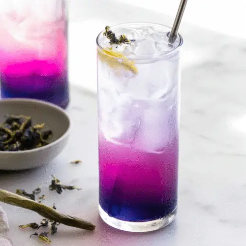 Vibrant butterfly pea lemonade in a tall glass, transitioning from blue to purple and yellow, garnished with fresh lemongrass and ginger slices for an herbal twist.