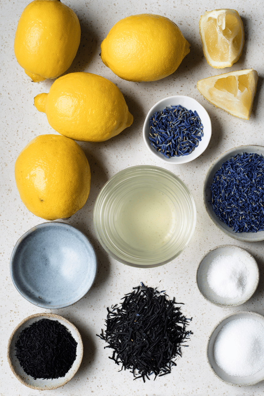A flat lay of fresh ingredients for butterfly pea lemonade, featuring dried butterfly pea flowers, sliced lemons, whole lemongrass stalks, and fresh ginger.