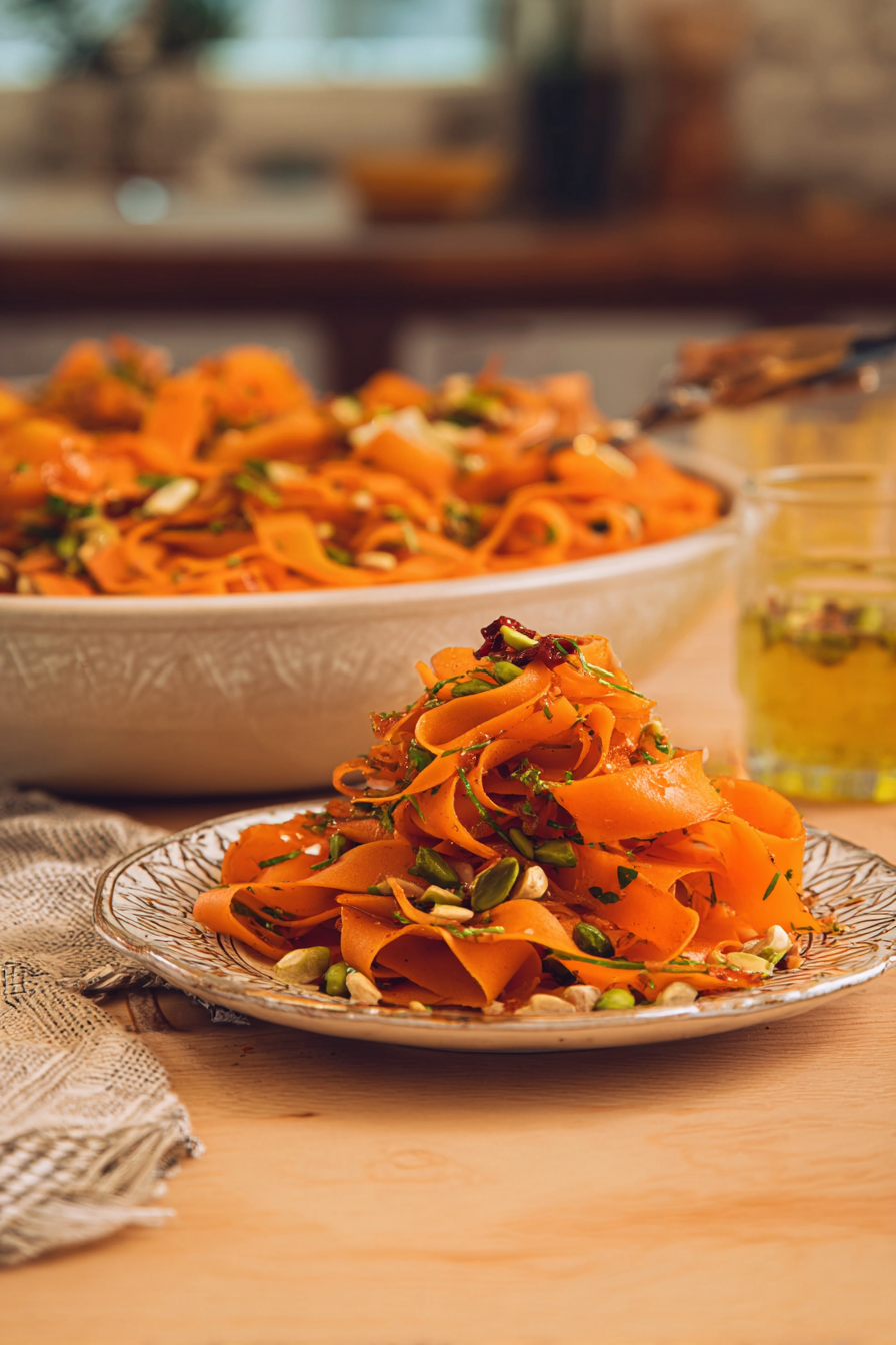 A vibrant serving of carrot ribbon salad with chili crisp placed on a wooden table, garnished with fresh cilantro and crushed pistachios.