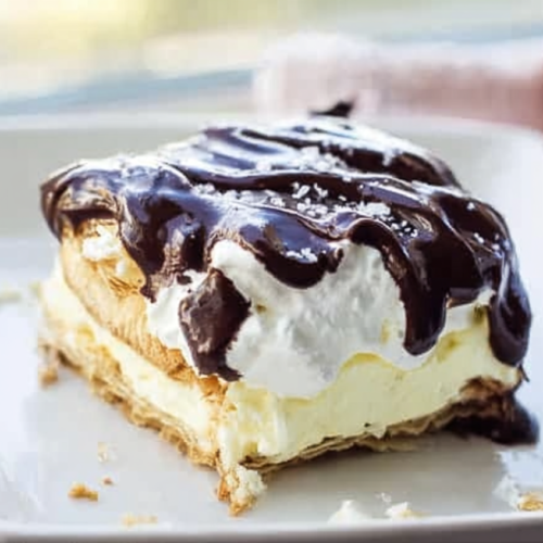 A luscious slice of Chocolate Eclair Cake with a creamy filling, drizzled chocolate ganache, and delicate sea salt flakes on top.