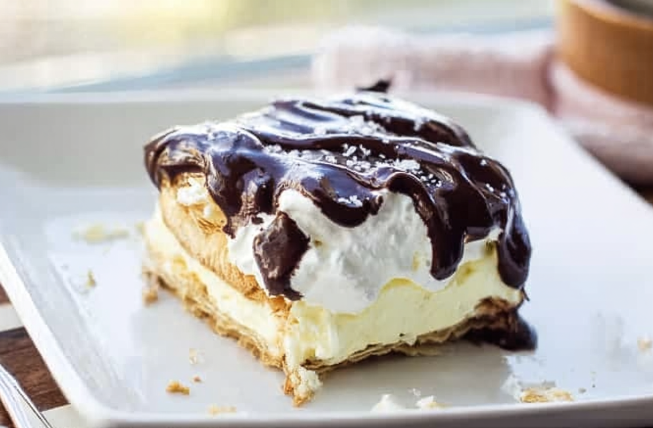 A luscious slice of Chocolate Eclair Cake with a creamy filling, drizzled chocolate ganache, and delicate sea salt flakes on top.