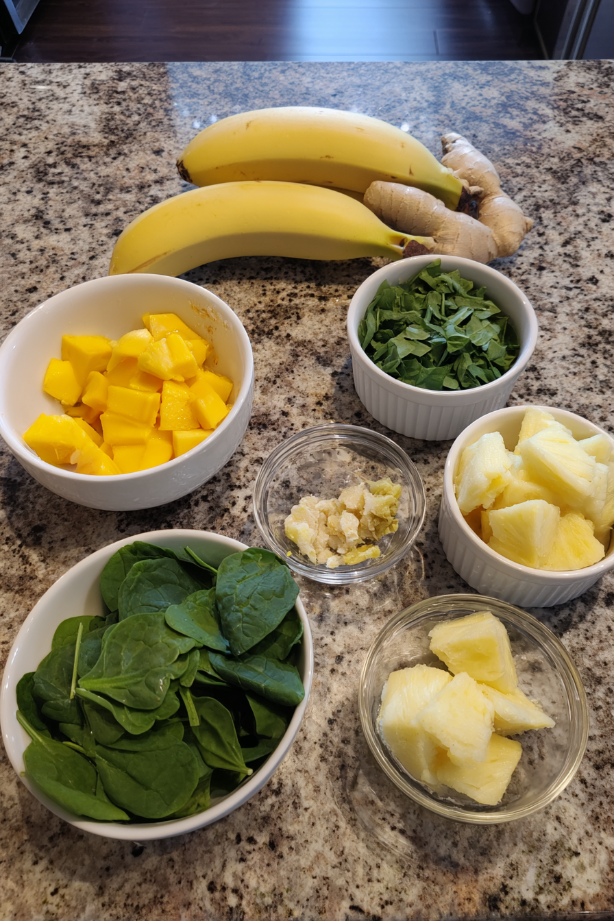 Raw ingredients for a Detox Island Green Smoothie, including fresh spinach, kale, mango, pineapple, banana, ginger, lime, and parsley, arranged on a flat surface.