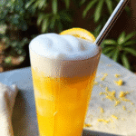 A tall glass of golden honey-ginger lemonade with cold foam, garnished with a lemon slice and fresh ginger on a light surface.