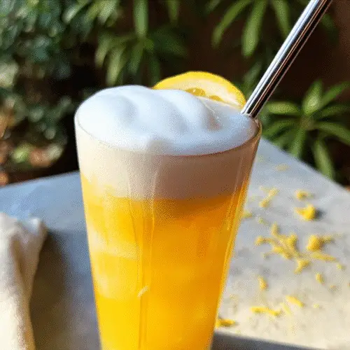 A tall glass of golden honey-ginger lemonade with cold foam, garnished with a lemon slice and fresh ginger on a light surface.