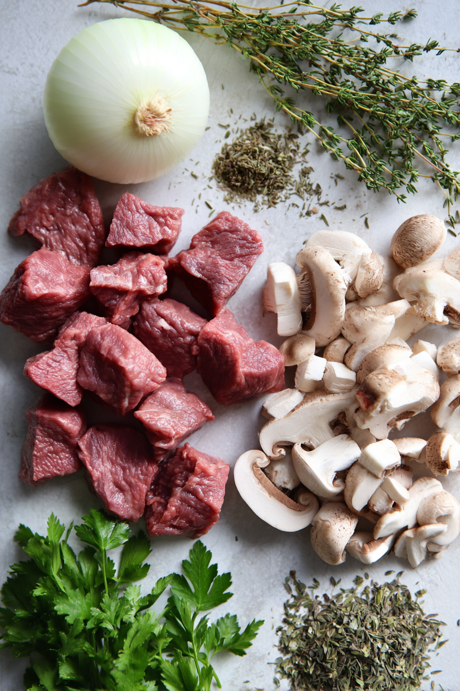 Overhead view of raw ingredients for beef tips crock pot recipes, including cubed beef stew meat, fresh mushrooms, onions, beef broth, and spices.