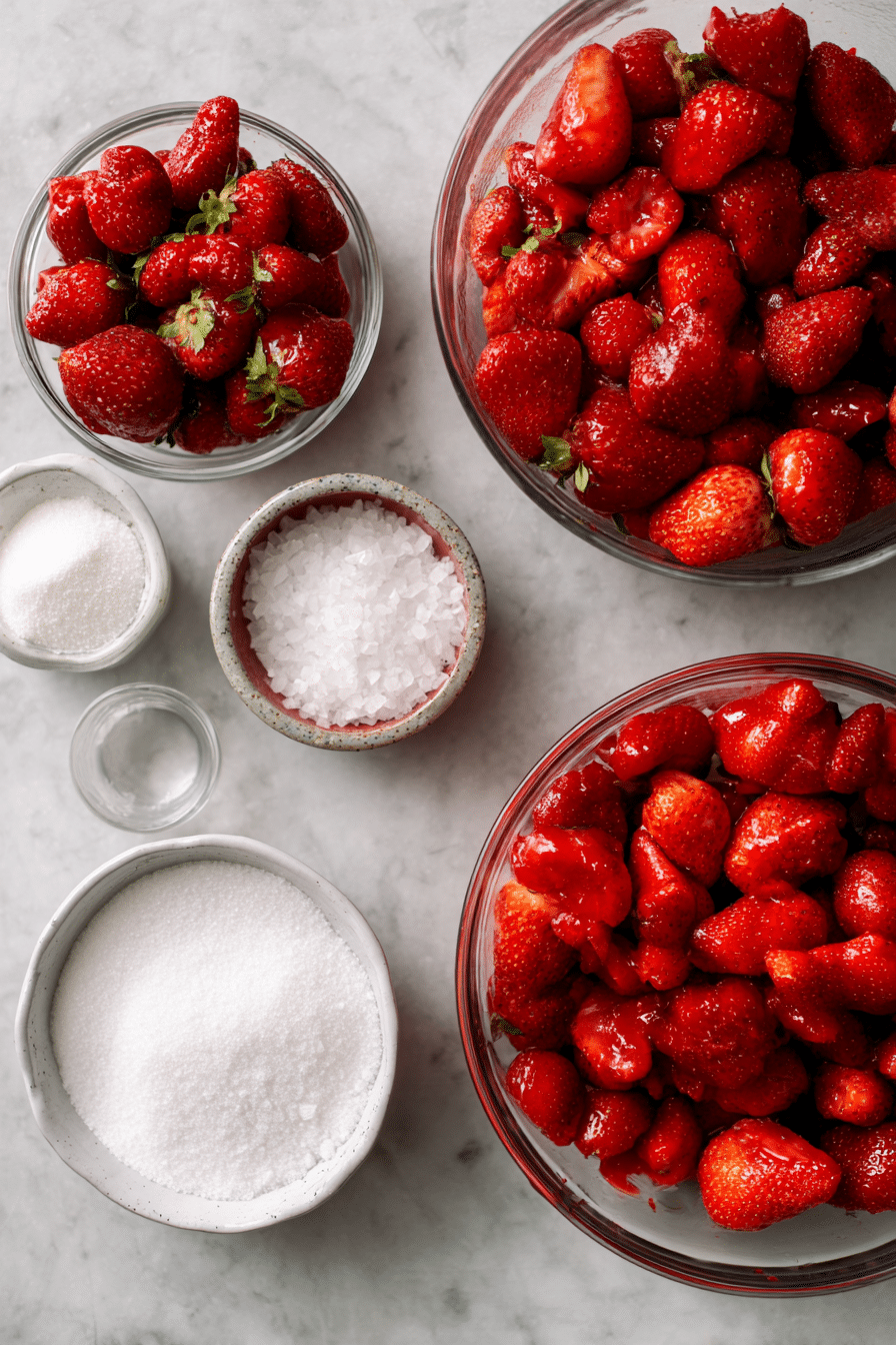 Overhead shot of fresh strawberries, sugar, a vanilla bean, and sea salt, laid out for a strawberry jam recipe.