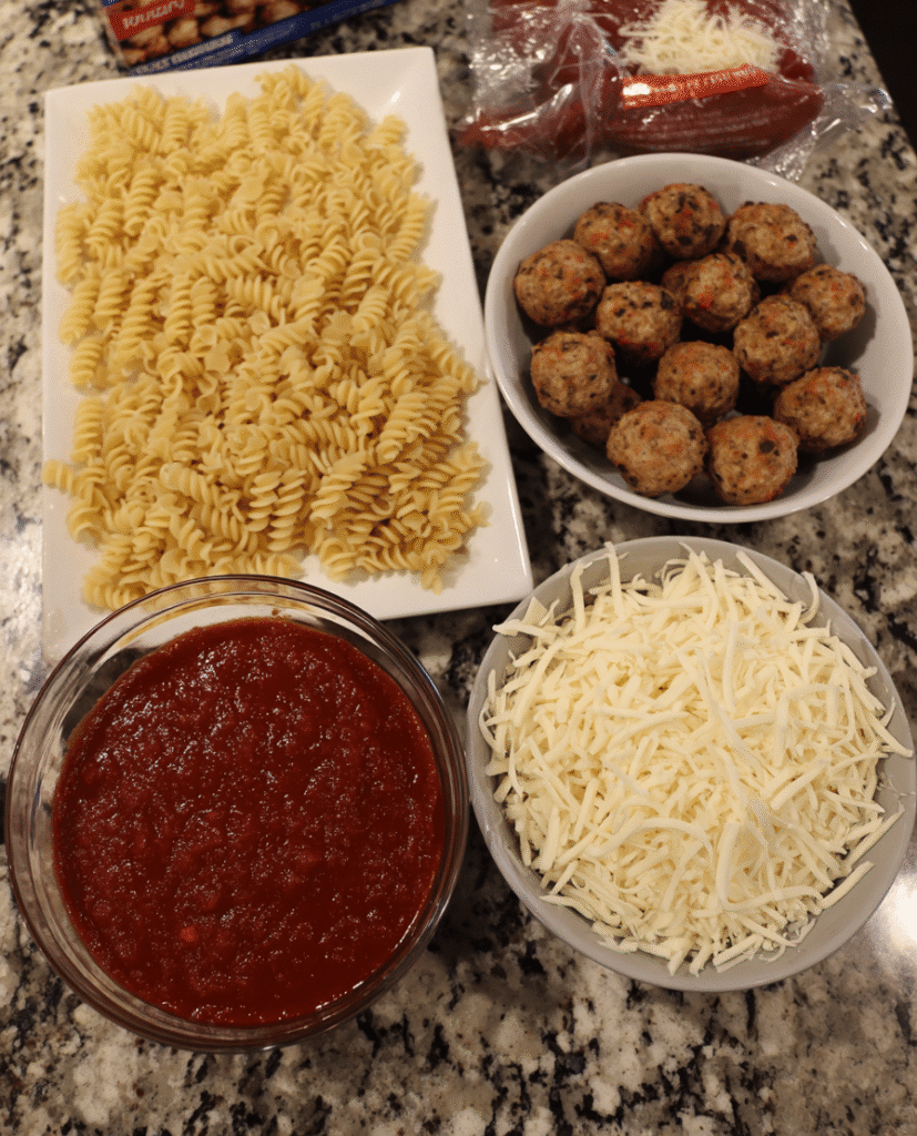 An overhead view of ingredients for Dump and Bake Meatball Casserole, including rotini pasta, frozen meatballs, pasta sauce, and shredded mozzarella cheese.