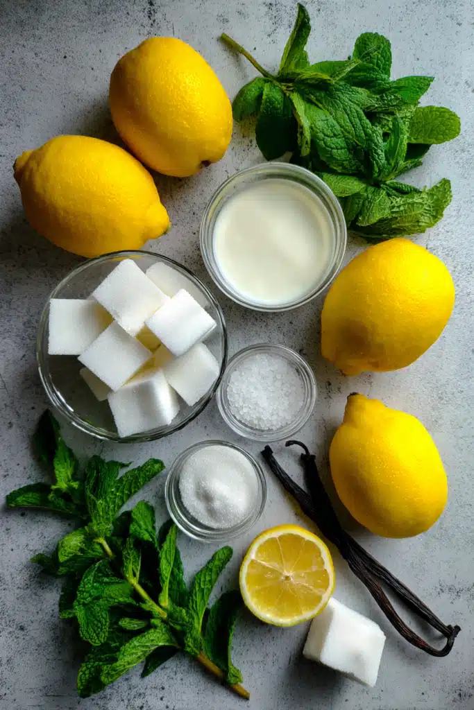 An overhead flat lay of fresh lemons, sliced ginger, honey, heavy cream, and a milk frother, ready for homemade lemonade with cold foam.