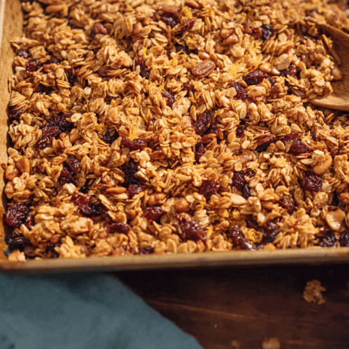 A close-up of a golden-brown granola recipe, rich with oats, nuts, and dried cranberries, infused with a bright orange essence.
