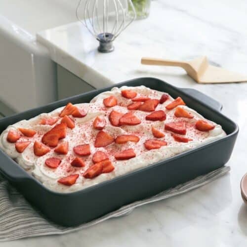 A slice of strawberry tres leches cake stands on a white plate, showcasing its moist texture and a delicate pink hue from the freeze-dried strawberry powder.