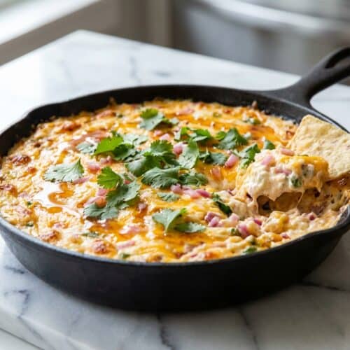 Creamy jalapeno popper dip in a skillet topped with melted cheese, crispy bacon, hot honey, and pickled red onions.