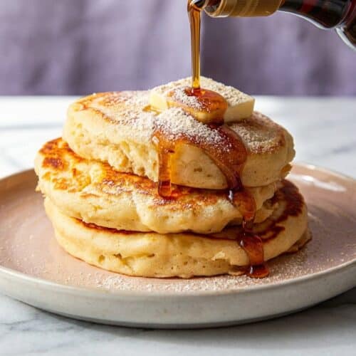A tall stack of fluffy pancakes with malted milk powder drizzled with maple syrup and topped with butter.