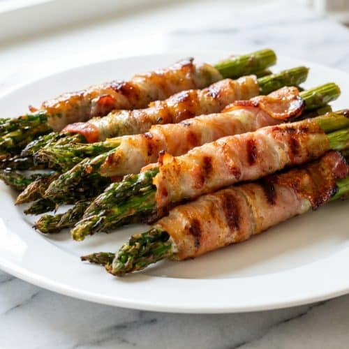 Bacon wrapped asparagus bundles coated in a glossy umami-maple glaze on a white serving platter.