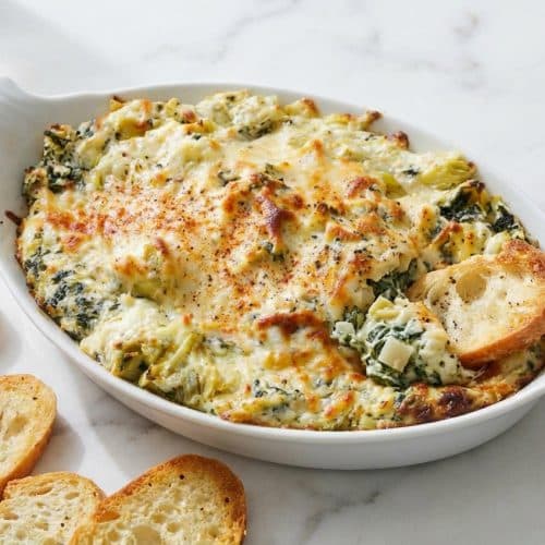 Bubbly spinach artichoke dip topped with smoked paprika and crunchy water chestnuts in a white ceramic baking dish.
