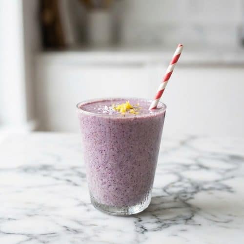 Frosty glass filled with a thick Blueberry Banana Protein Smoothie, garnished with lemon zest and sea salt to highlight the brightening twist.