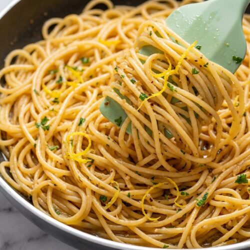 Twirled garlic butter pasta with golden brown butter bits and a fresh lemon zest finish on a white plate.