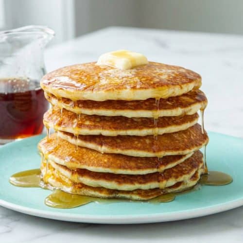 Stack of fluffy buttermilk pancakes with golden browned butter edges and a syrup drizzle in a cast iron skillet.