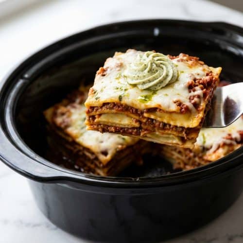 A scoop of crockpot ravioli lasagna in a white bowl, showing layers of pasta, meat sauce, and a creamy ricotta-pesto swirl on top.