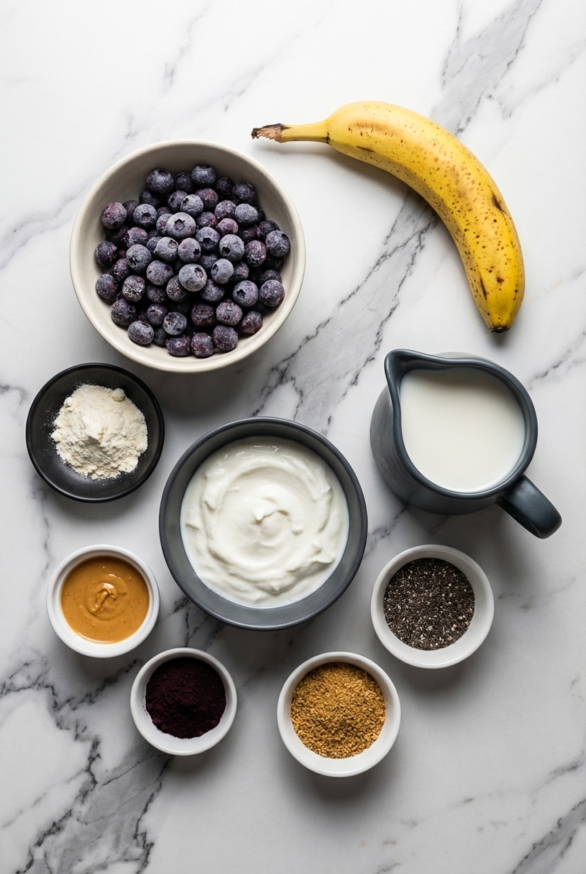 Overhead view of frozen blueberries, ripe banana, whey protein isolate powder, and lemon slices arranged for a Blueberry Banana Protein Smoothie.