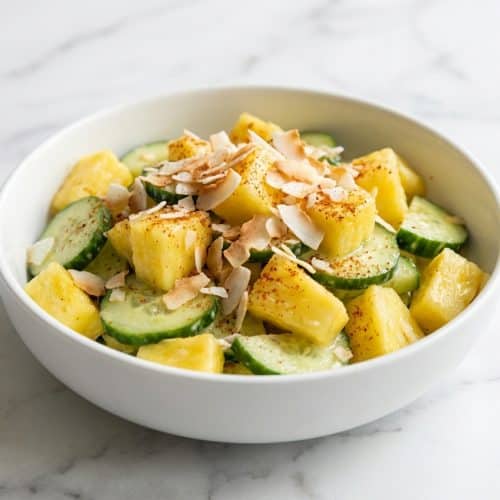 Pineapple cucumber salad topped with toasted coconut flakes and a dusting of smoked paprika in a white bowl.