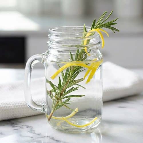Steaming glass mug of our rosemary tea recipe, featuring bruised fresh sprigs and water infused with bright lemon zest.
