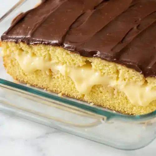 Boston Cream Poke Cake slice showing the vanilla bean milk soak and glossy, sea salt topped chocolate ganache.