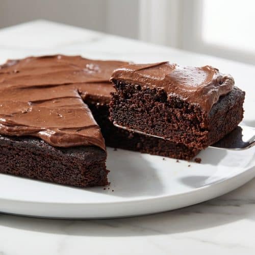 A slice of chocolate mayonnaise cake on a white plate, topped with a thick, glossy layer of brown butter chocolate frosting.