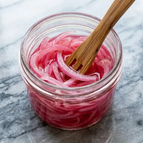 Vibrant pink quick pickled onions in a jar with visible toasted peppercorns and a hint of earthy sumac.