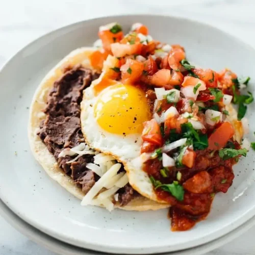 A plate of authentic huevos rancheros with two sunny-side-up eggs over corn tortillas, covered in red salsa and showing a perfectly runny