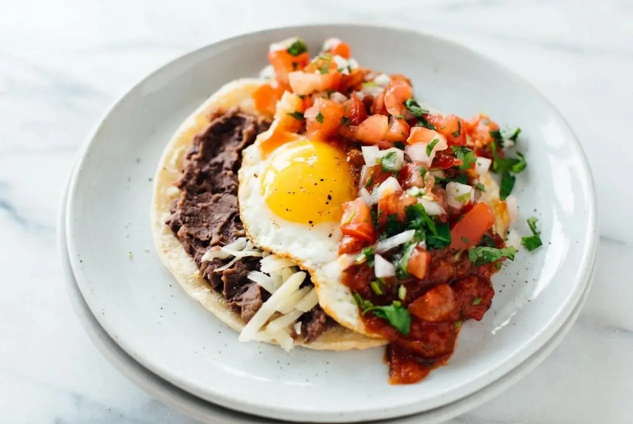 A plate of authentic huevos rancheros with two sunny-side-up eggs over corn tortillas, covered in red salsa and showing a perfectly runny