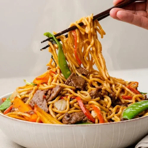 A large white bowl of beef lo mein recipe with tender beef slices, chewy noodles, and crisp vegetables all coated in a glossy savory sauce.