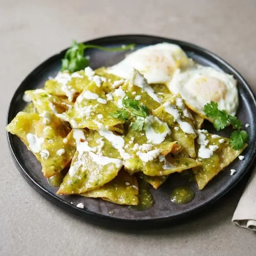 A skillet of chilaquiles verdes coated in tangy green salsa, topped with crumbled queso fresco, crema, and fresh cilantro.