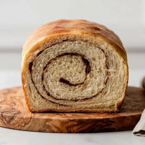 A loaf of homemade cinnamon swirl bread, partially sliced to reveal a gooey cinnamon sugar spiral and soft, fluffy crumb.
