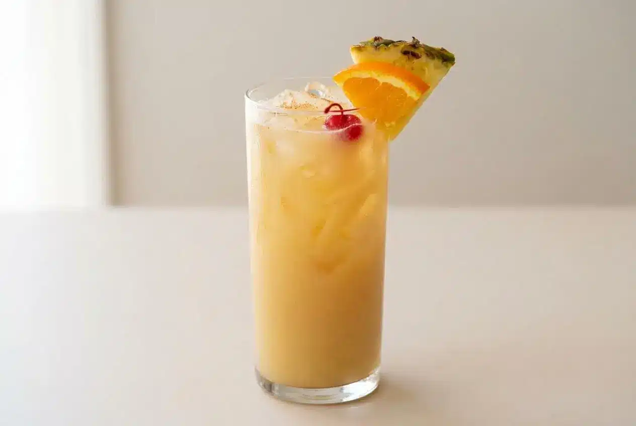 A classic Painkiller Cocktail in a highball glass, showing a creamy frothy top, and garnished with a pineapple wedge and a cherry.