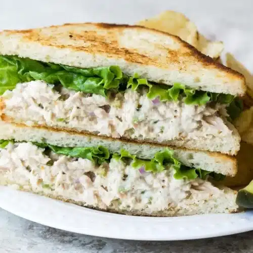 A white bowl filled with a creamy and classic tuna salad recipe, showing flakes of tuna and bits of celery and red onion.