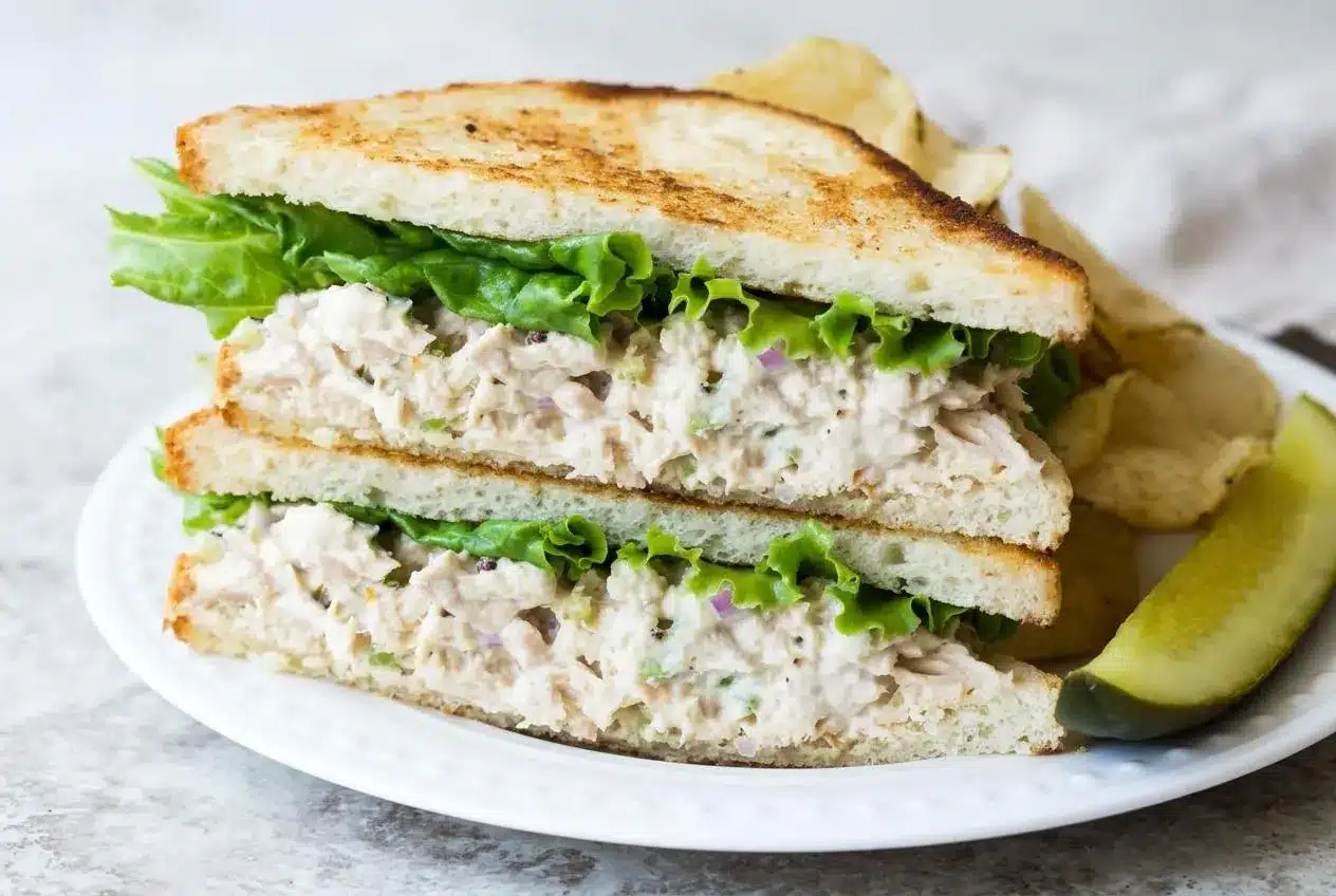 A white bowl filled with a creamy and classic tuna salad recipe, showing flakes of tuna and bits of celery and red onion.