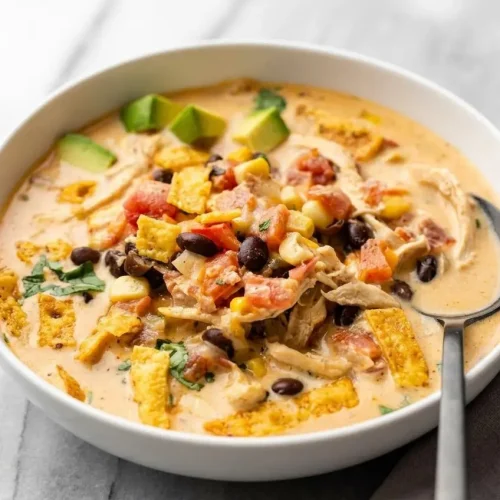 A white bowl of creamy chicken taco soup topped with shredded cheese, fresh cilantro, and crispy tortilla strips.