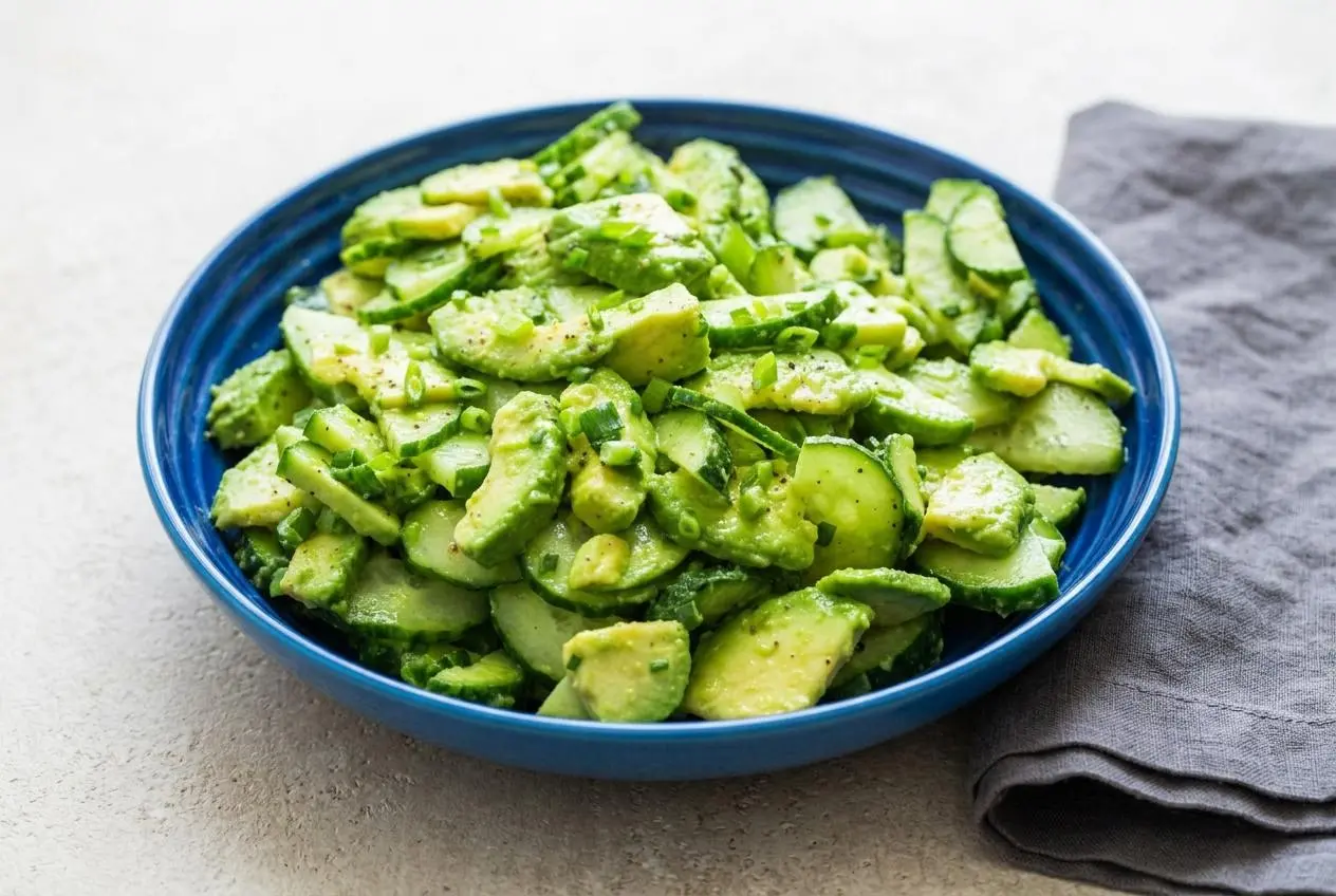 A large white bowl of cucumber avocado salad, showing creamy sliced avocado and crisp cucumber with a light, glossy dressing.