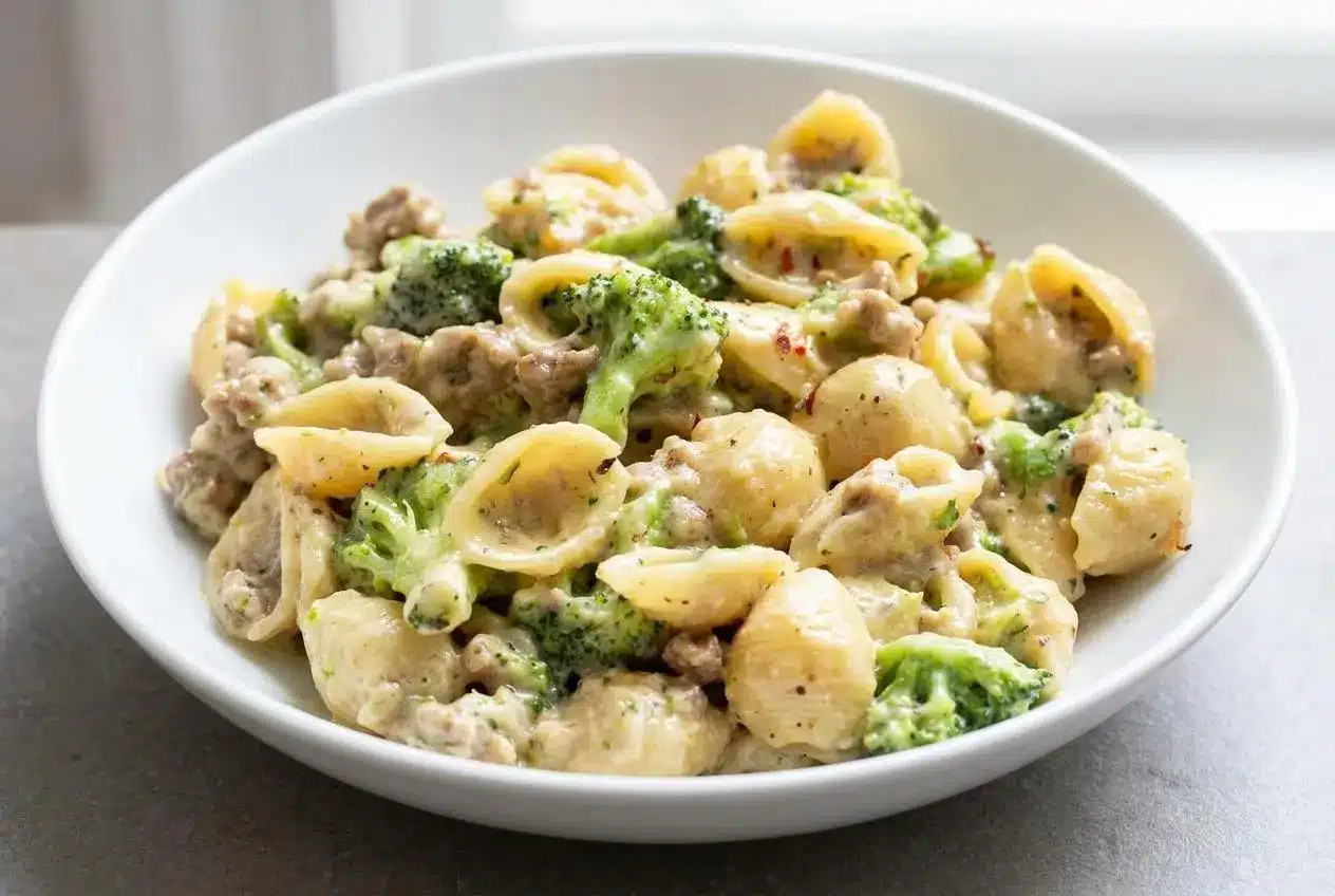 A white bowl of creamy sausage and broccoli pasta, showing shell pasta coated in a rich sauce with savory sausage and bright green broccoli