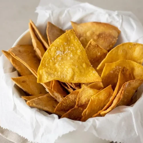 A dark gray bowl filled with crispy, golden Air Fryer Tortilla Chips, with some scattered on a dark slate surface.