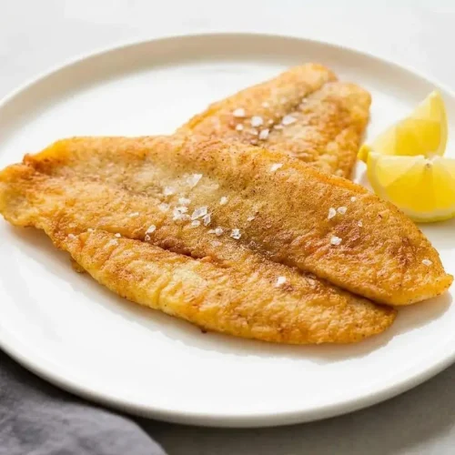 Two crispy golden fillets from a fried fish recipe, served on a dark plate with a lemon wedge and fresh parsley.