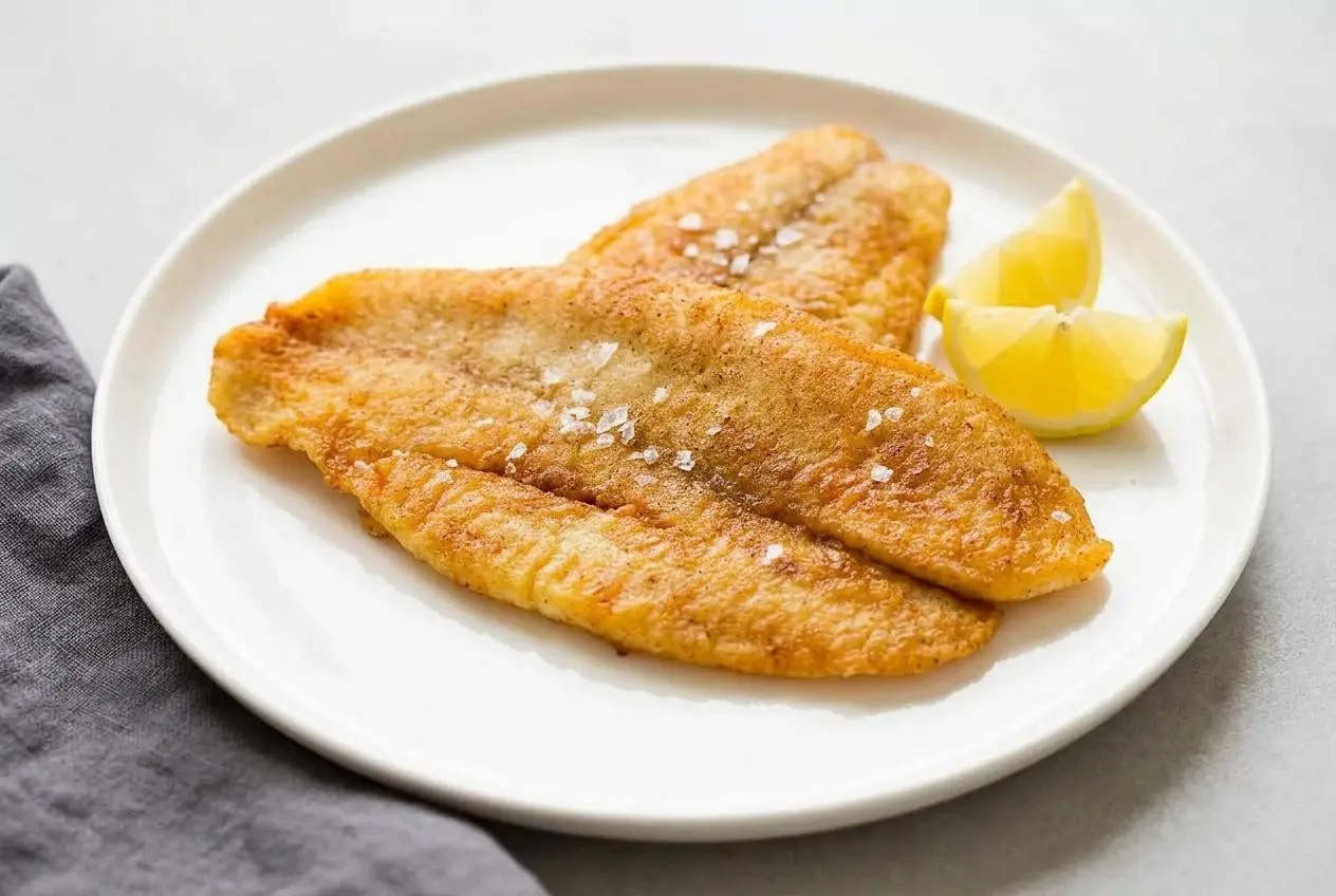 Two crispy golden fillets from a fried fish recipe, served on a dark plate with a lemon wedge and fresh parsley.