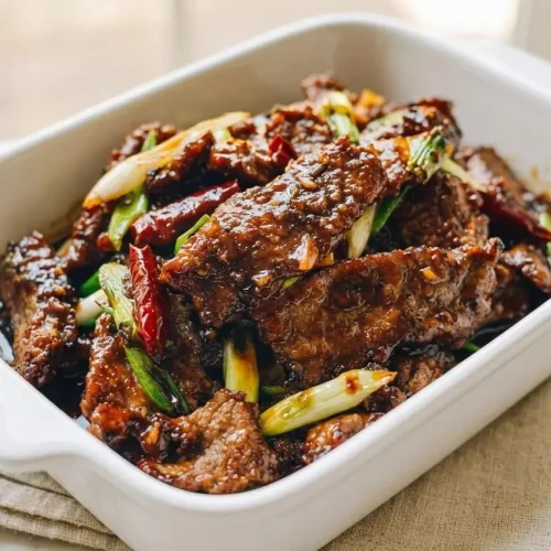 A rustic ceramic bowl filled with the finished Mongolian beef recipe, coated in a sticky, glossy sauce and garnished with green onions.