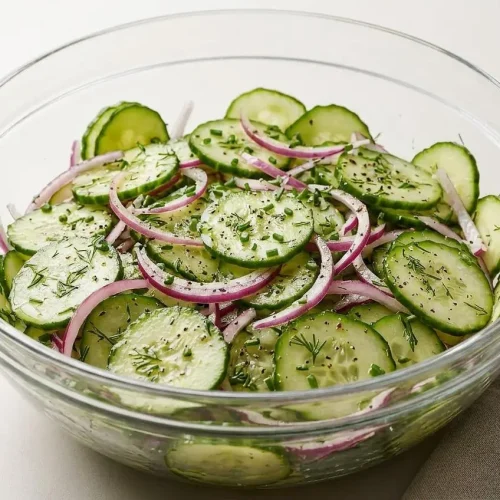 A white serving bowl filled with Cucumber Dill Salad, showing thin, crisp cucumber and red onion slices coated in a light vinaigrette.