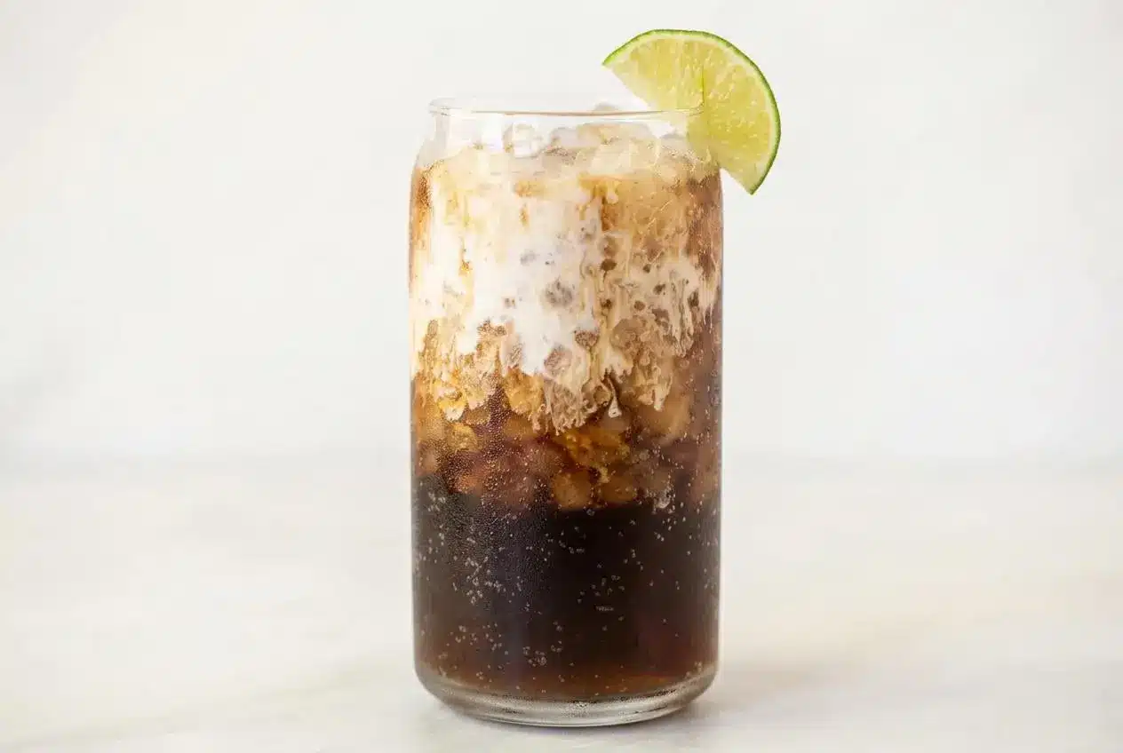 A tall glass of a creamy dirty dr pepper recipe, filled with pebble ice and garnished with a fresh lime wedge and a black straw.