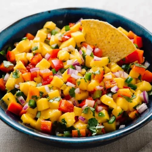 A white bowl filled with fresh, chunky mango salsa, showing diced mango, red bell pepper, red onion, and cilantro.