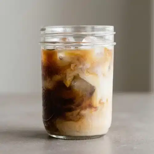 A tall glass filled with a creamy, layered iced latte recipe, topped with milk foam and served on a dark surface.